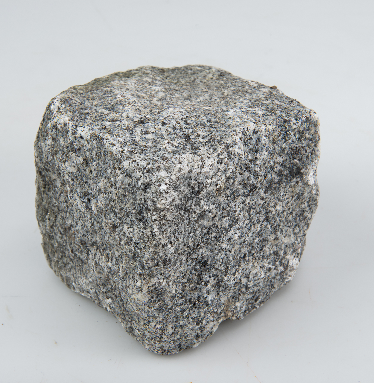En ganska grovt huggen gatsten av granit. Stenen är nästan kubisk. Den är grå i flera nyanser.
