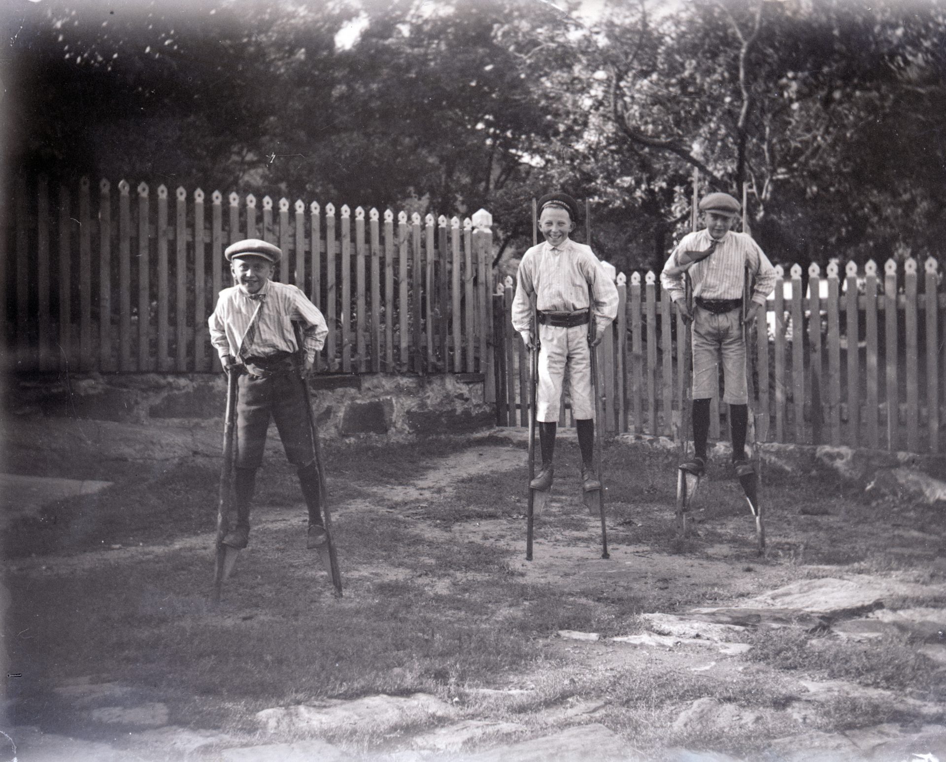 Svartvit bild av tre pojkar som går på styltor. I bakgrunden ett staket.