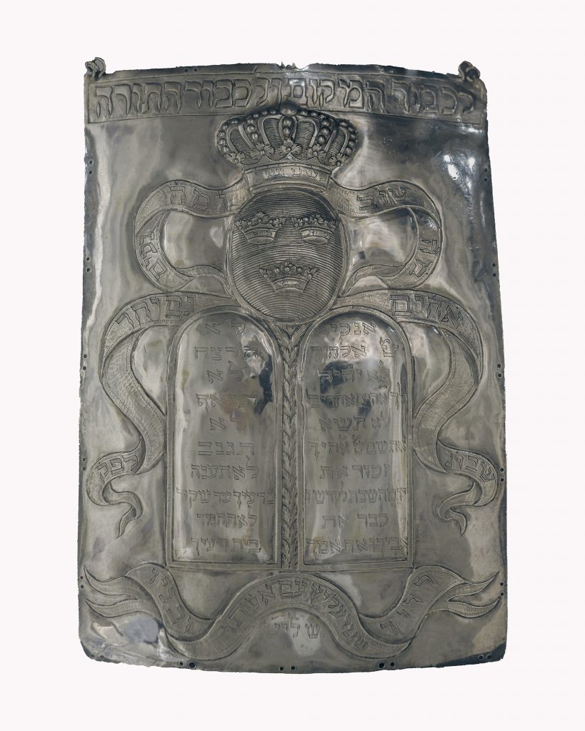 En silversköld med relief av tre kronor, en slinga med skrift på hebreiska och två fält med text.