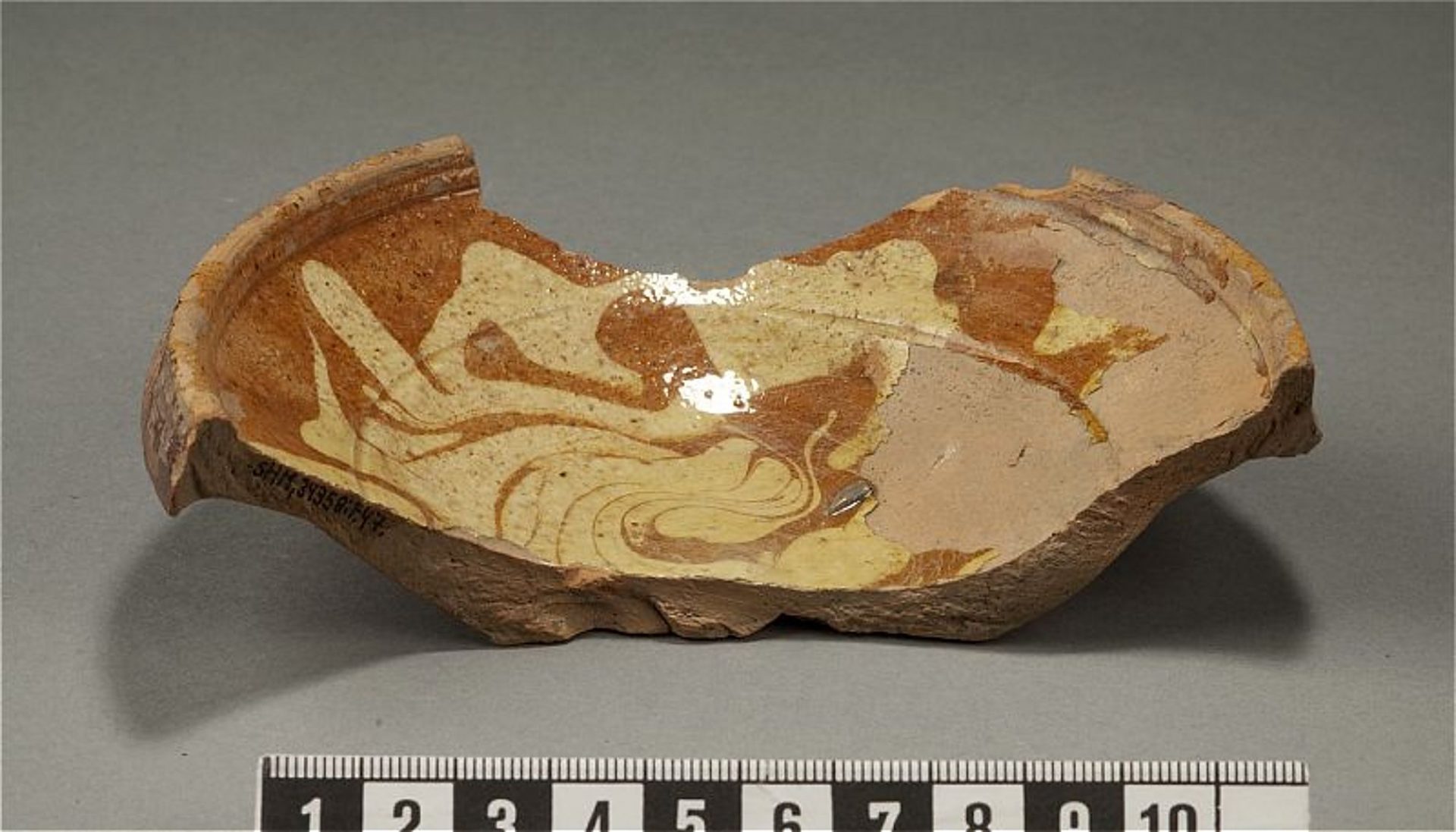 Skärvan av en keramikskål är ldekorerad i rödbrunt och vitt och glaserad. Skålen är vid och har en tjockare kant.
