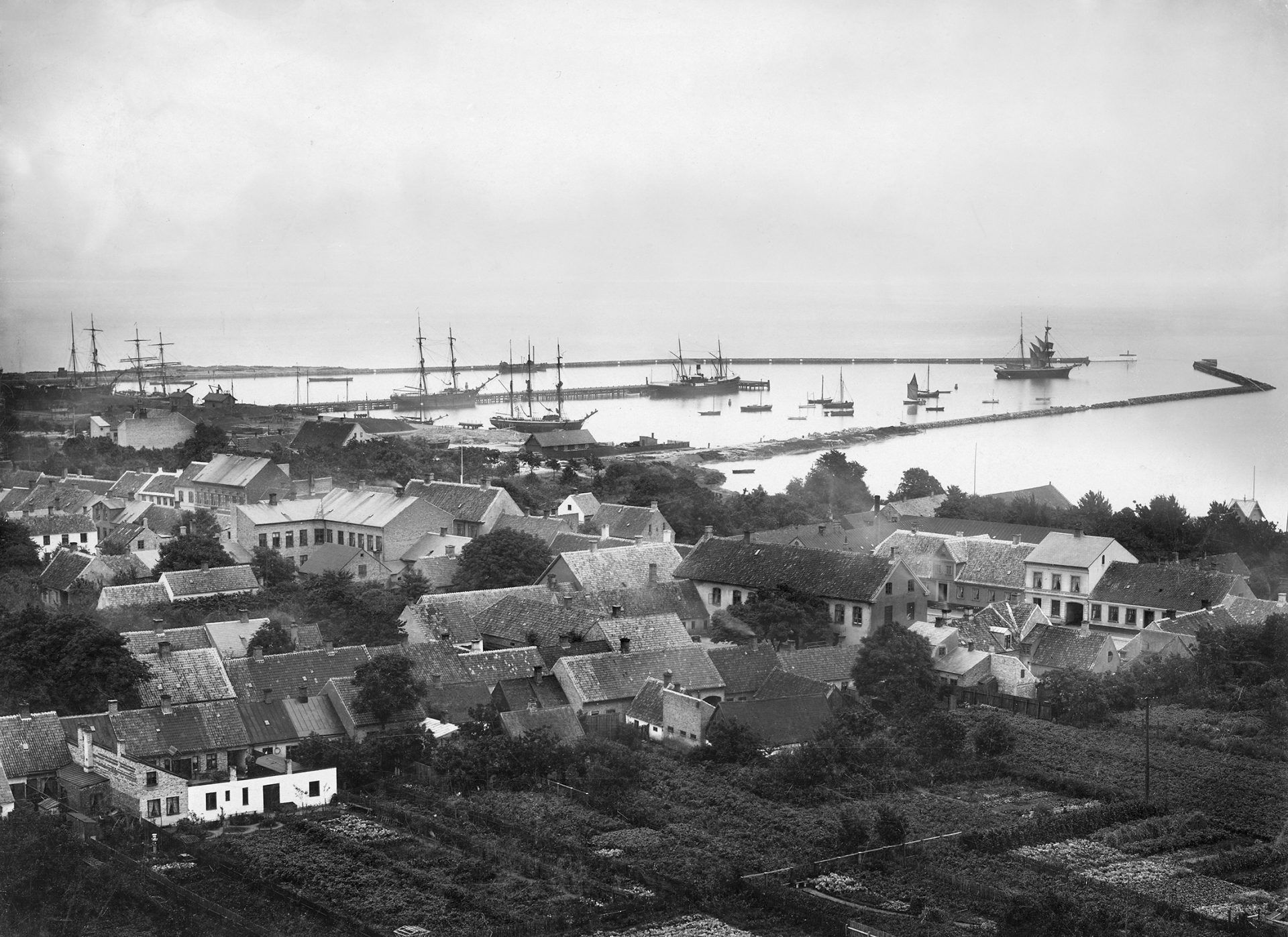 Ett gammalt fotografi som visar en mängd små hus med en hamn med segelfartyg i bakgrunden.