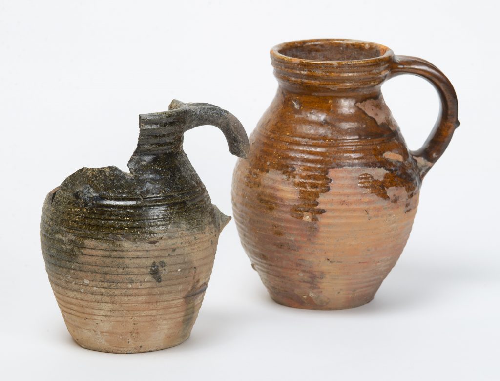 Två keramikkannor, en är trasig med svart glasyr. Den andra är hel och har brun glasyr.