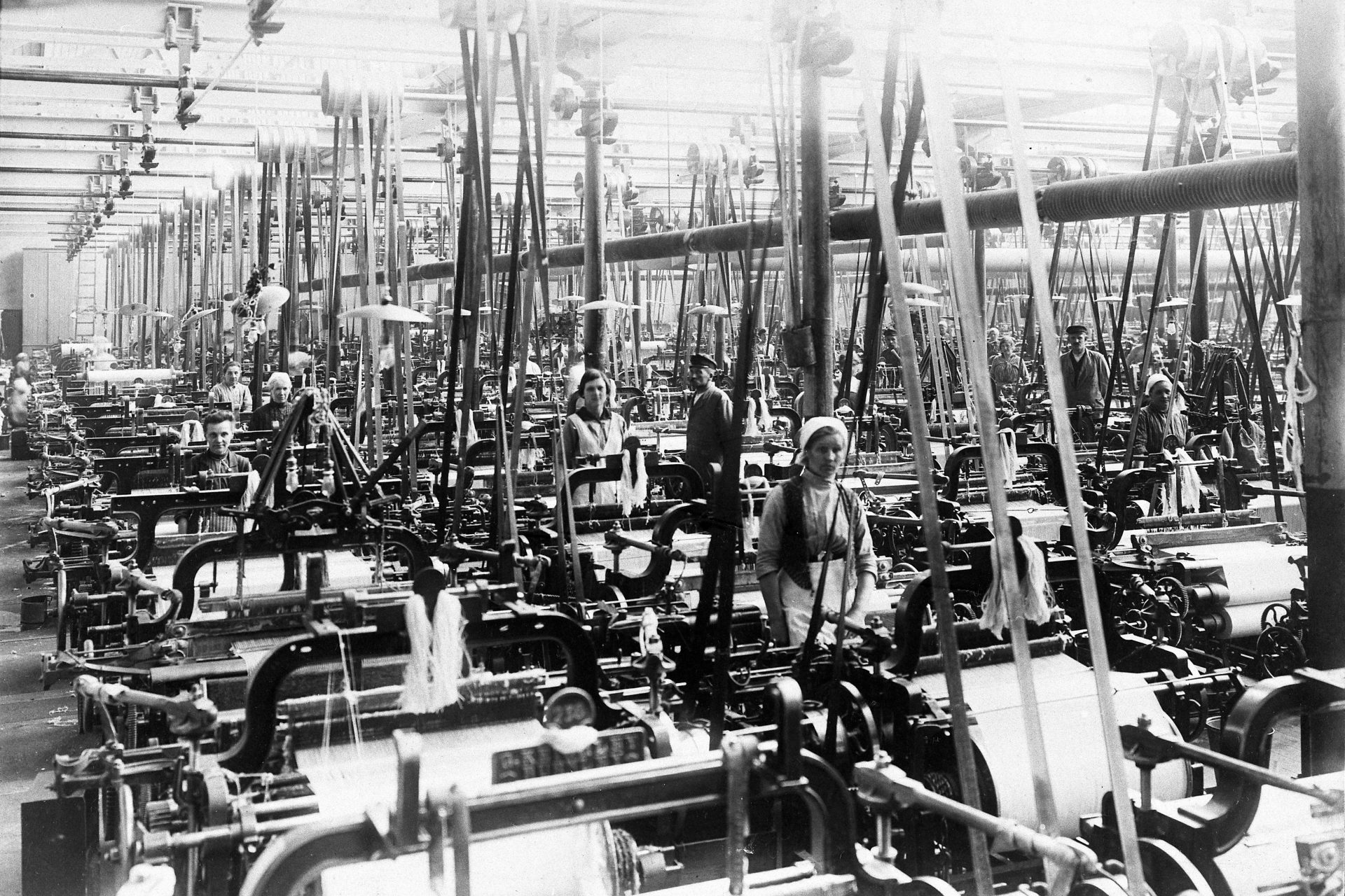 En stor maskinhall med hundratals vävmaskiner. Bland maskinerna står några kvinnor och män.