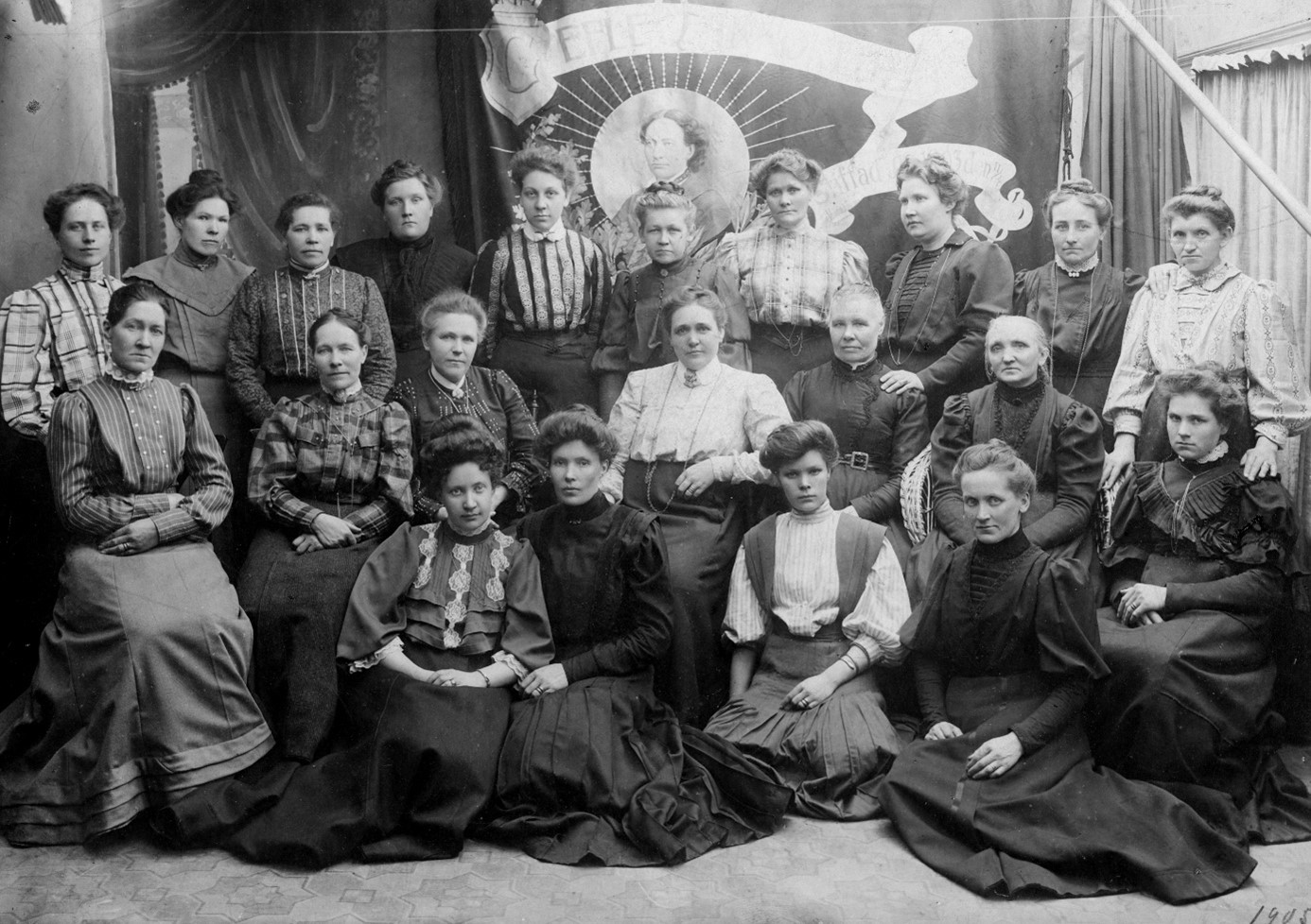 Arton kvinnor sittandes framför en fana.