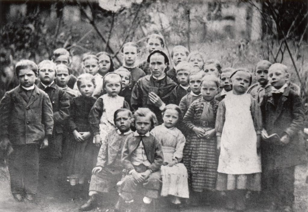 Svartvitt foto av en grupp barn. I mitten sitter en vuxen kvinna.