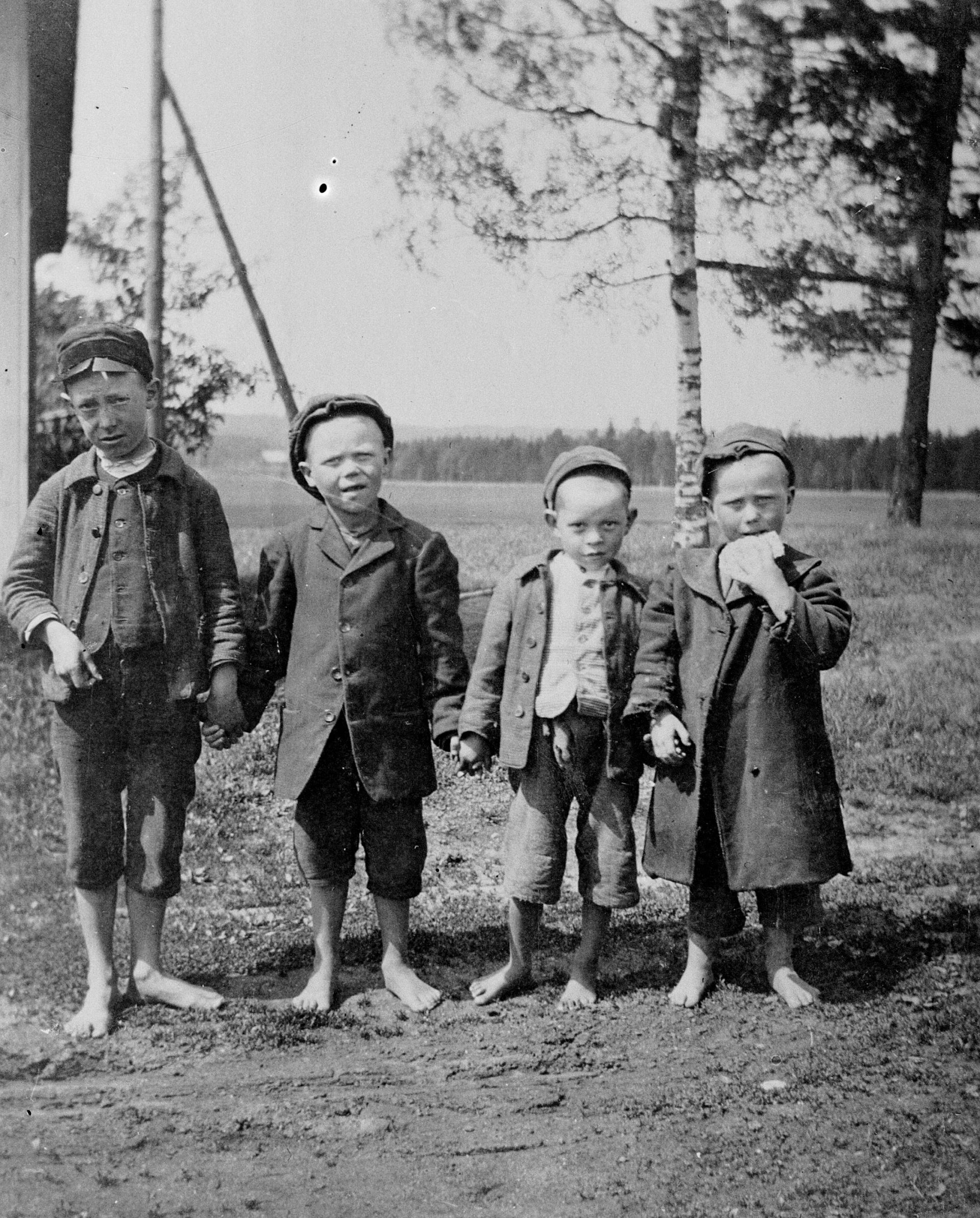 Fyra små pojkar med kläder men utan skor. I bakgrunden en husknut och några träd.