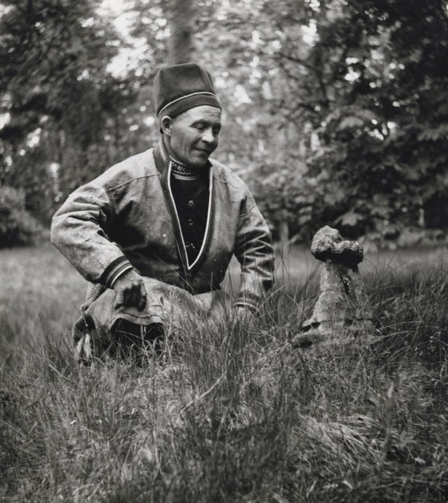 En samisk man sitter på huk framför en sten (sejte)