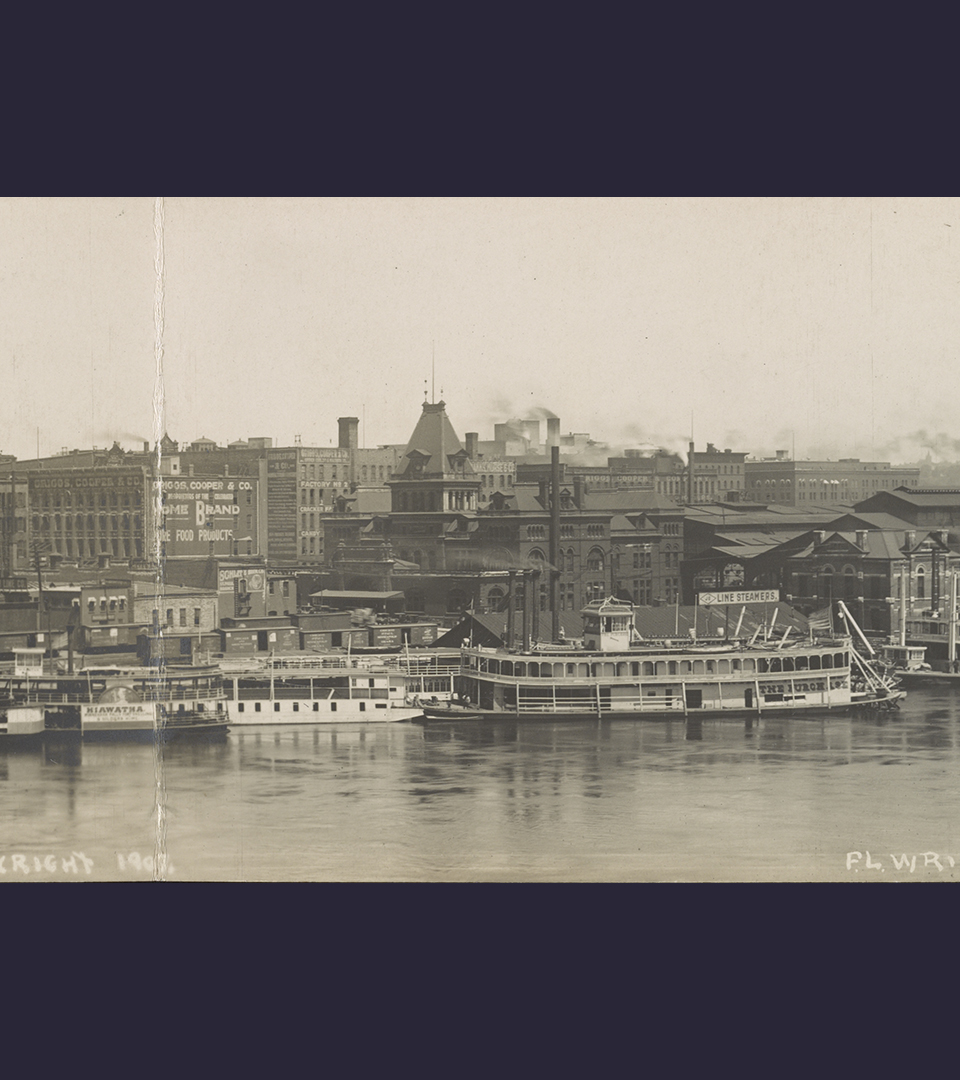 Svartvitt foto av en stad. I förgrunden vatten och flera större båtar som ligger förtöjda vid kajen.