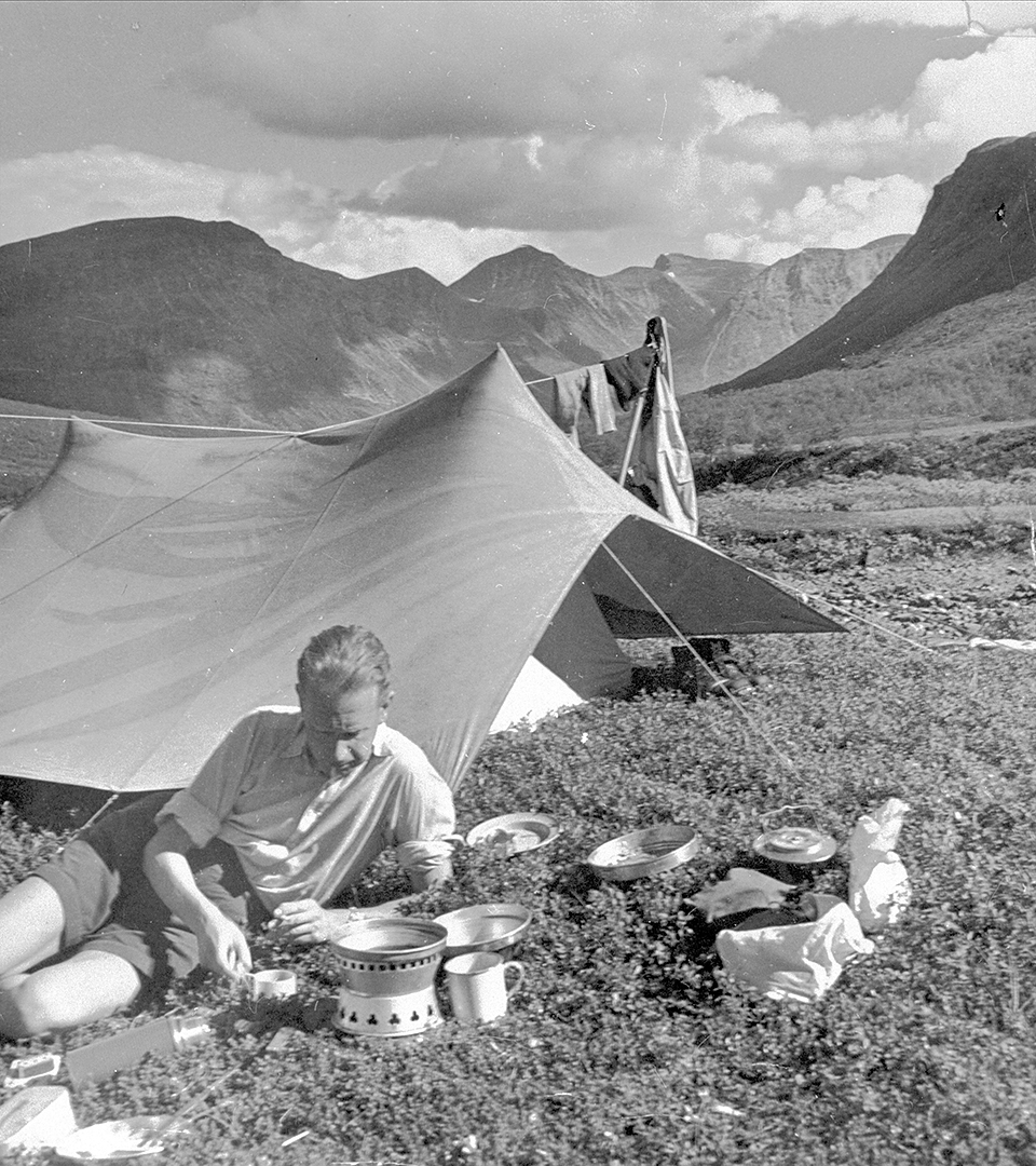 En man halvligger på marken, framför ett tält. I bakgrunden ser man berg och en molning himmel.