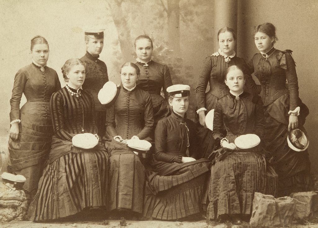 Svartvitt foto av nio unga kvinnor i mörka klänningar. Några sitter och några står. Två har studentmössor på huvudet och de andra håller mössorna i handen.