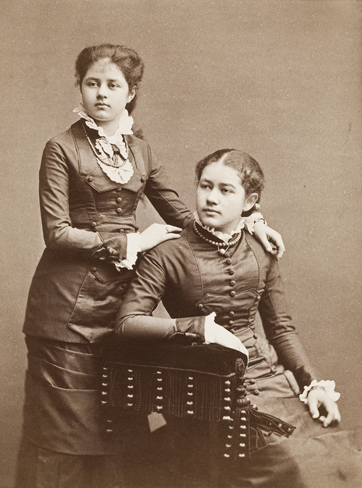 Svartvitt foto av två flickor i övre tonåren. En sitter och den andra står bakom henne.