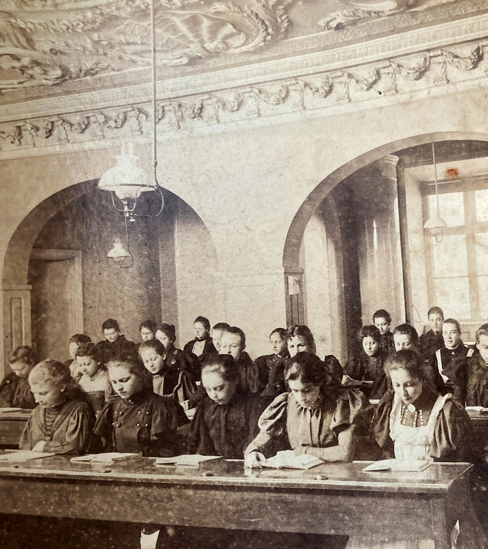 Svartvitt foto som visar flickor som sitter i skolbänkar i en stor, vacker skolsal.