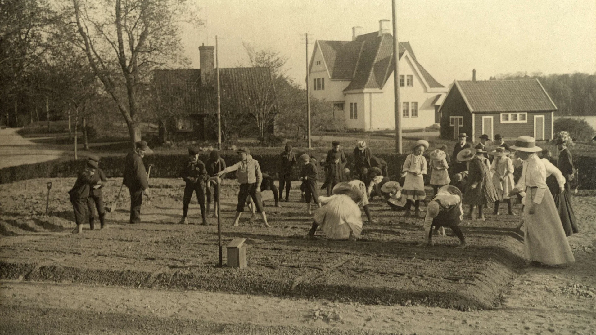 Svartvitt foto av en massa barn med trädgårdsredskap i händerna. De står i ett grönsaksland.