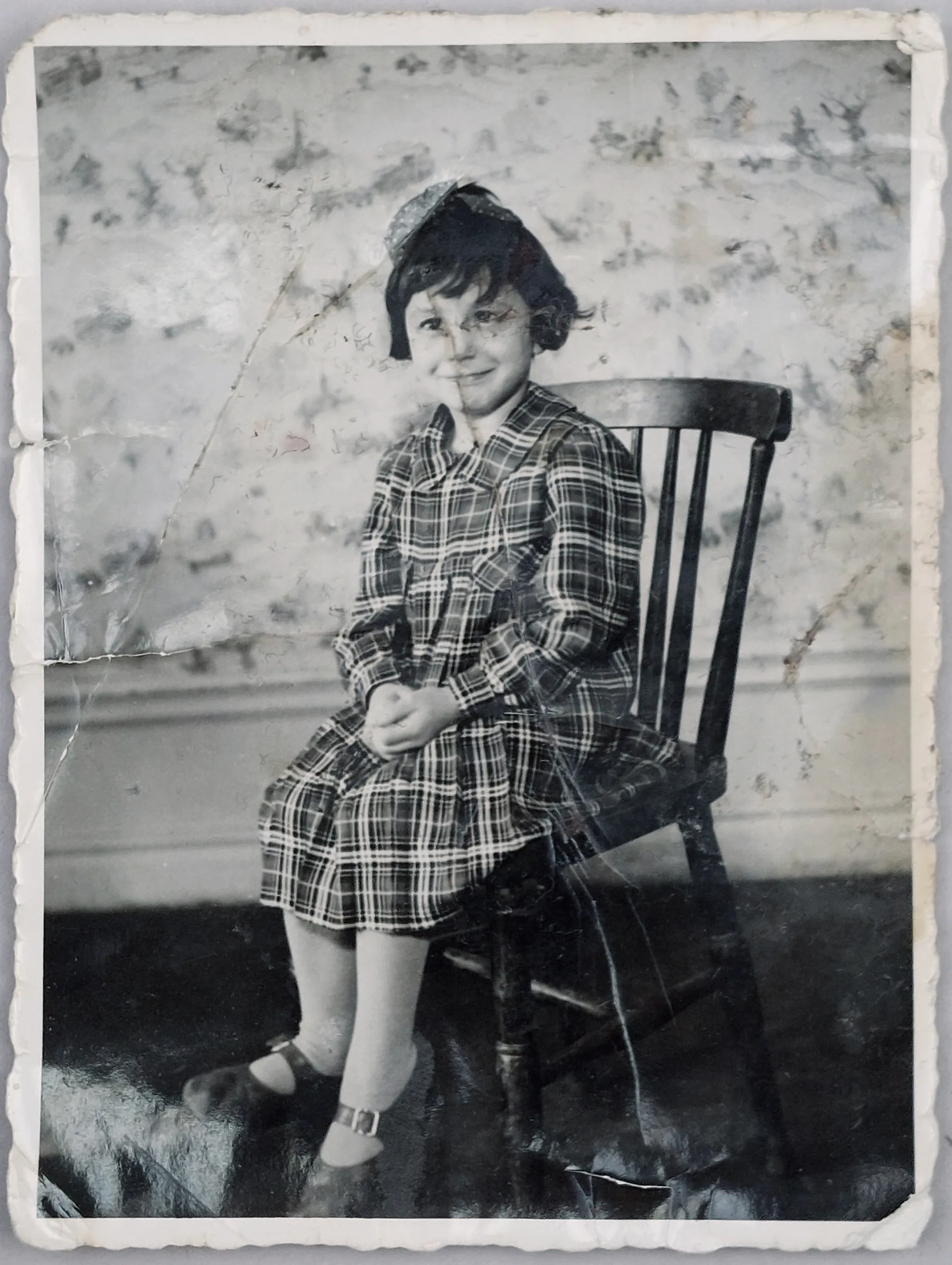 Porträtt av en ung flicka som sitter på en stol. Hon är iklädd en rutig klänning.