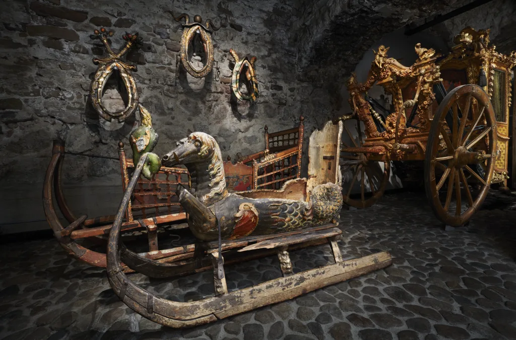 träsläde med en sittplats med ryggstöd. Dekorerad med två djurhuvuden och fiskfjäll. I bakgrunden ytterligare en släde och en del av en förgylld vagn